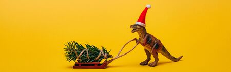Panoramic shot of toy dinosaur in santa hat with pine on sleigh on yellow backgroundの写真素材