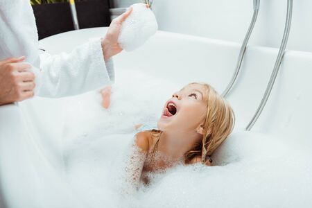 excited kid looking at hand of mother in bath foam in bathroomの写真素材