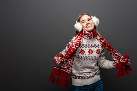 attractive smiling woman in christmas sweater, scarf, mittens and earmuffs, isolated on grey