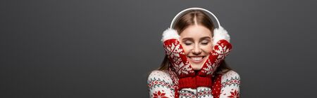 panoramic shot of happy girl with closed eyes in christmas sweater, scarf, mittens and earmuffs,...