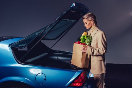 side view of attractive and stylish woman in suit standing near retro car and holding paper bag with foodの写真素材