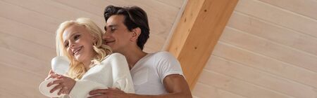 Low angle view of smiling man hugging girlfriend with coffee at home, panoramic shot