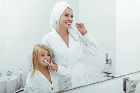 happy mother and daughter in bathrobes brushing teeth near mirror
