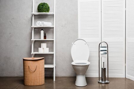 interior of modern bathroom with toilet bowl near rack with cosmetics, towels, toilet paper, laundry basket and toilet brushの写真素材