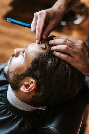 selective focus of barber holding sharp razor while doing haircut to man 