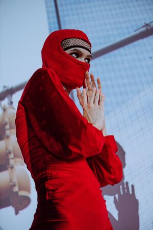 low angle view of stylish woman in red dress and balaclava on city background 