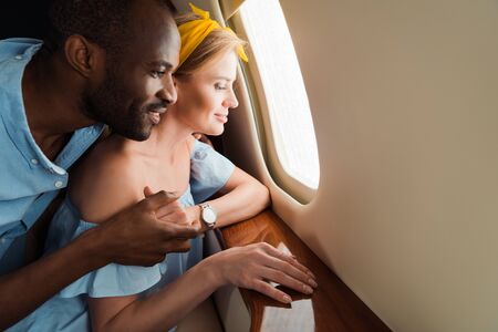 happy african american man and cheerful woman looking at airplane window in private planeの写真素材