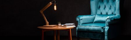 elegant velour blue armchair with pillow near wooden table and lamp isolated on black, panoramic shotの写真素材