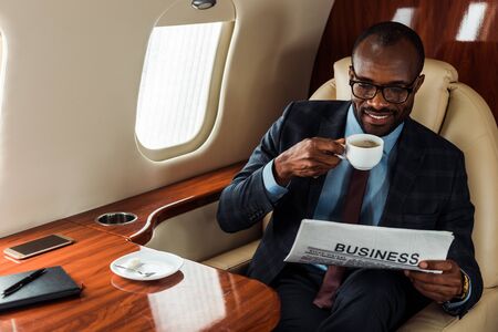 cheerful african american businessman in glasses holding business newspaper and cup in private plane
