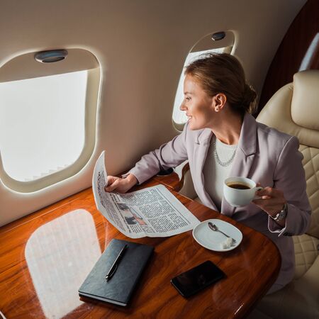 happy businesswoman holding cup and business newspaper near smartphone with blank screen in private...