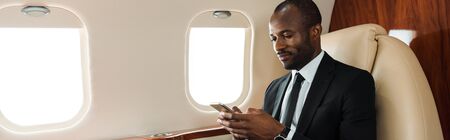 panoramic shot of handsome african american businessman using smartphone in private jet