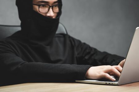 selective focus of asian hacker with obscured face using laptop and sitting at table