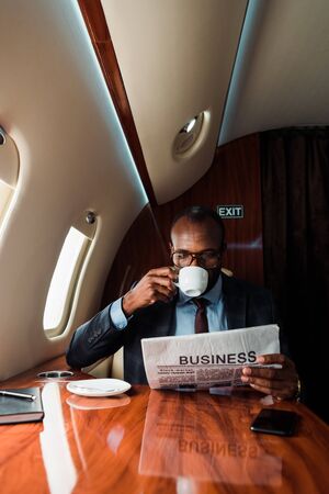 African american businessman in glasses reading business newspaper while drinking coffee in private planeの写真素材