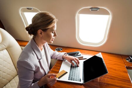 attractive businesswoman using laptop with blank screen and holding credit card in private plane