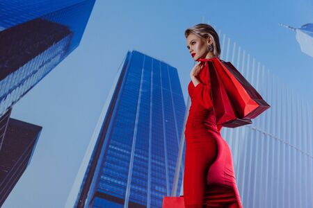low angle view of attractive and stylish woman in red dress holding shopping bags on city backgroundの写真素材