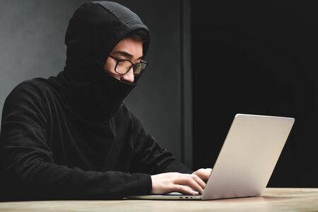 asian hacker with obscured face using laptop and sitting at tableの写真素材
