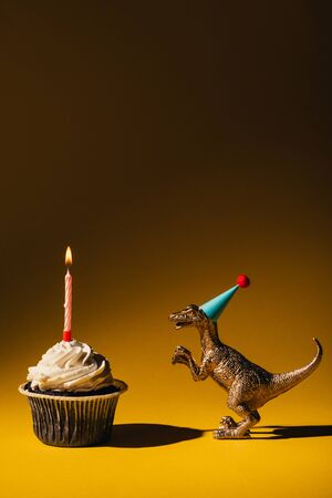 Toy dinosaur in party cap beside cupcake with burning candle on brown backgroundの写真素材