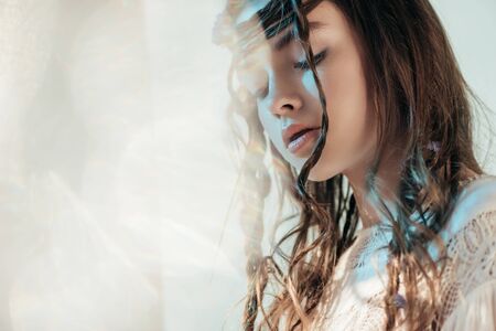 tender young woman with braids in hairstyle posing in white boho dress on grey with lens flaresの写真素材