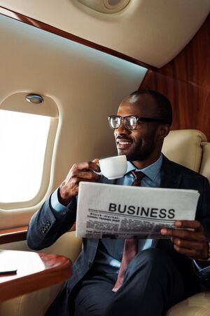 happy african american businessman in glasses holding business newspaper and cup in private plane