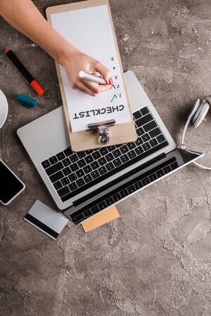 top view of woman holding marker pen near checklist, gadgets, credit cards and headphones,...