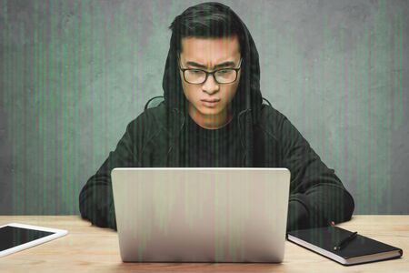 asian hacker in glasses sitting at table, using laptop and surrounded with illustration