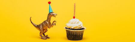 Panoramic shot of toy dinosaur in party cap and cupcake with candle on yellow backgroundの写真素材