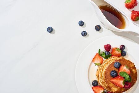 top view of delicious pancakes with maple syrup, blueberries and strawberries on plate on marble...