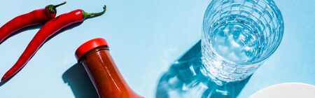 Top view of glass of water with chili sauce and chili peppers on blue background, panoramic shot