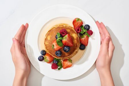 partial view of woman holding plate with delicious pancakes with honey, blueberries and strawberries on marble white surfaceの写真素材
