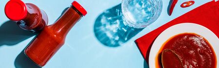 Top view of ketchup in plate and bottles beside chili pepper and glass of water on blue surface,...