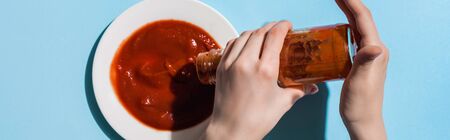 Cropped view of woman pouring tasty ketchup from bottle to plate on blue surface, panoramic shotの写真素材
