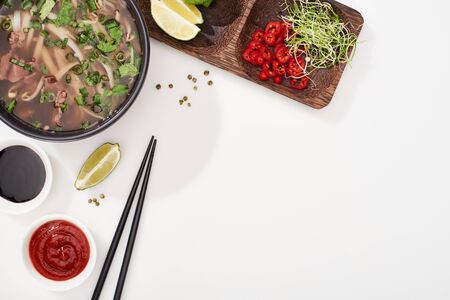 top view of pho in bowl near chopsticks, lime, chili and soy sauces and coriander on white...
