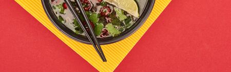 top view of pho in bowl with chopsticks, lime, chili and coriander on red and yellow background,...