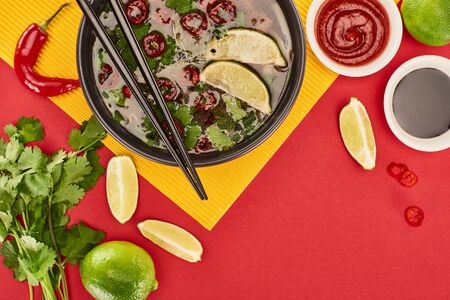 top view of pho in bowl with chopsticks near sauces, lime, chili and coriander on red and yellow backgroundの写真素材