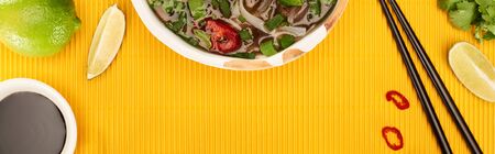 top view of pho in bowl near chopsticks, lime, soy sauce and coriander on yellow textured...