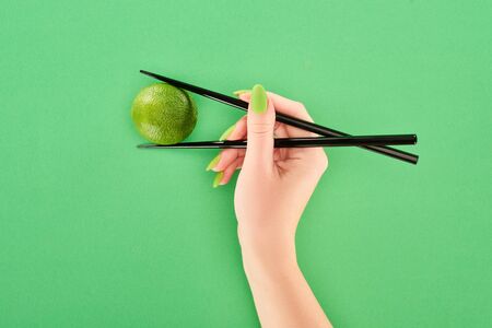 cropped view of woman holding whole lime with chopsticks on green background