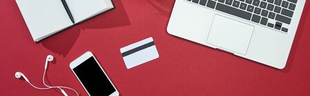top view of credit card on red background with smartphone, laptop, earphones, notebook, panoramic...