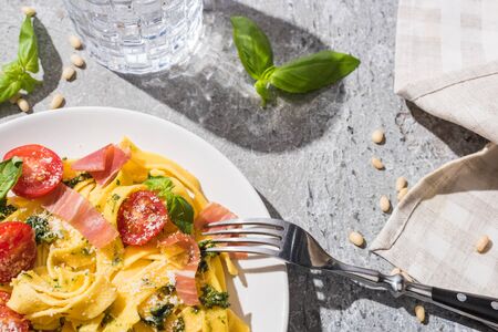 top view of tasty Pappardelle with tomatoes, pesto and prosciutto with fork near water and pine nuts on grey surfaceの写真素材