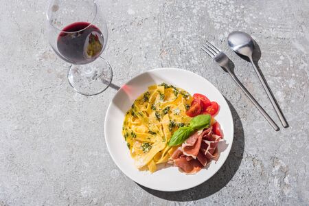 top view of tasty Pappardelle with tomatoes, pesto and prosciutto near red wine and cutlery on grey surfaceの写真素材