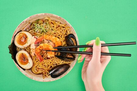 cropped view of woman eating spicy seafood ramen with chopsticks on green surface