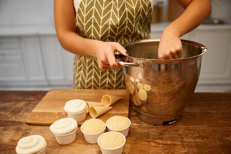 Cropped view of confectioner making cream in bowl beside fresh cupcakes and waffle cones table