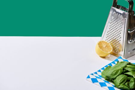 lemon, basil leaves and grater on white table isolated on green