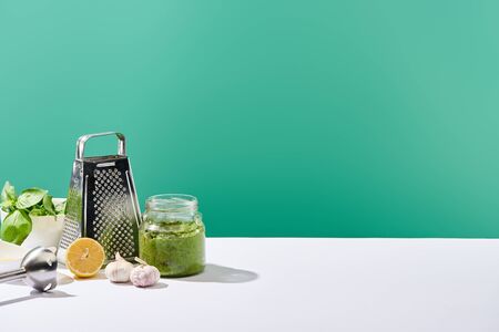 pesto sauce in jar near ingredients, blender and grater on white table isolated on green