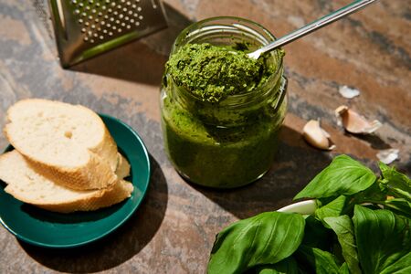 baguette slices, pesto sauce in jar, grater and fresh ingredients on stone surface