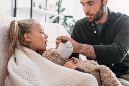attentive father giving medicines to sick daughter holding teddy bearの写真素材