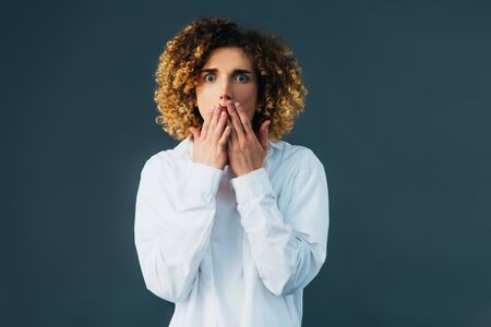 shocked stylish curly teenager in total white outfit covering mouth with hands isolated on green