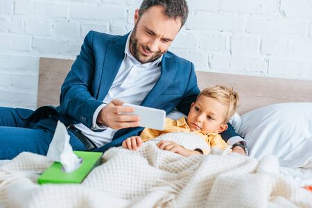 smiling father showing smartphone to sick son lying in bedの写真素材