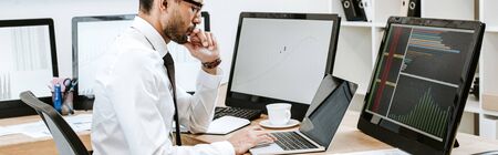 panoramic shot of bi-racial trader using laptop and sitting at tableの写真素材
