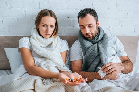 sick woman holding handful of pills while sitting in bed near diseased husbandの写真素材