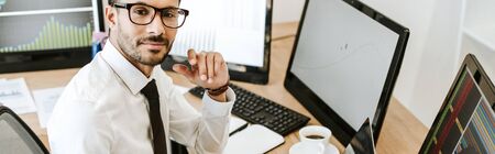 panoramic shot of smiling bi-racial trader sitting near computers and looking at cameraの写真素材